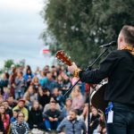 Artist med gitarr på utomhusscen framför publik en sommarkväll